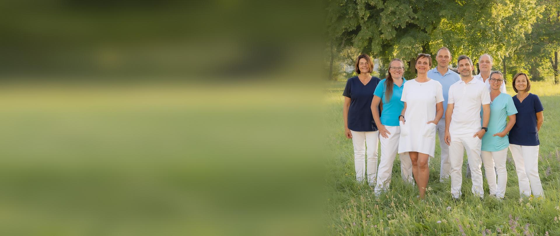 Das Ortho-Vital-Team – Ärztin, Ärzte, Therapeuten und Assistentinnen – steht gemeinsam auf einer sommerlichen Wiese vor Bäumen