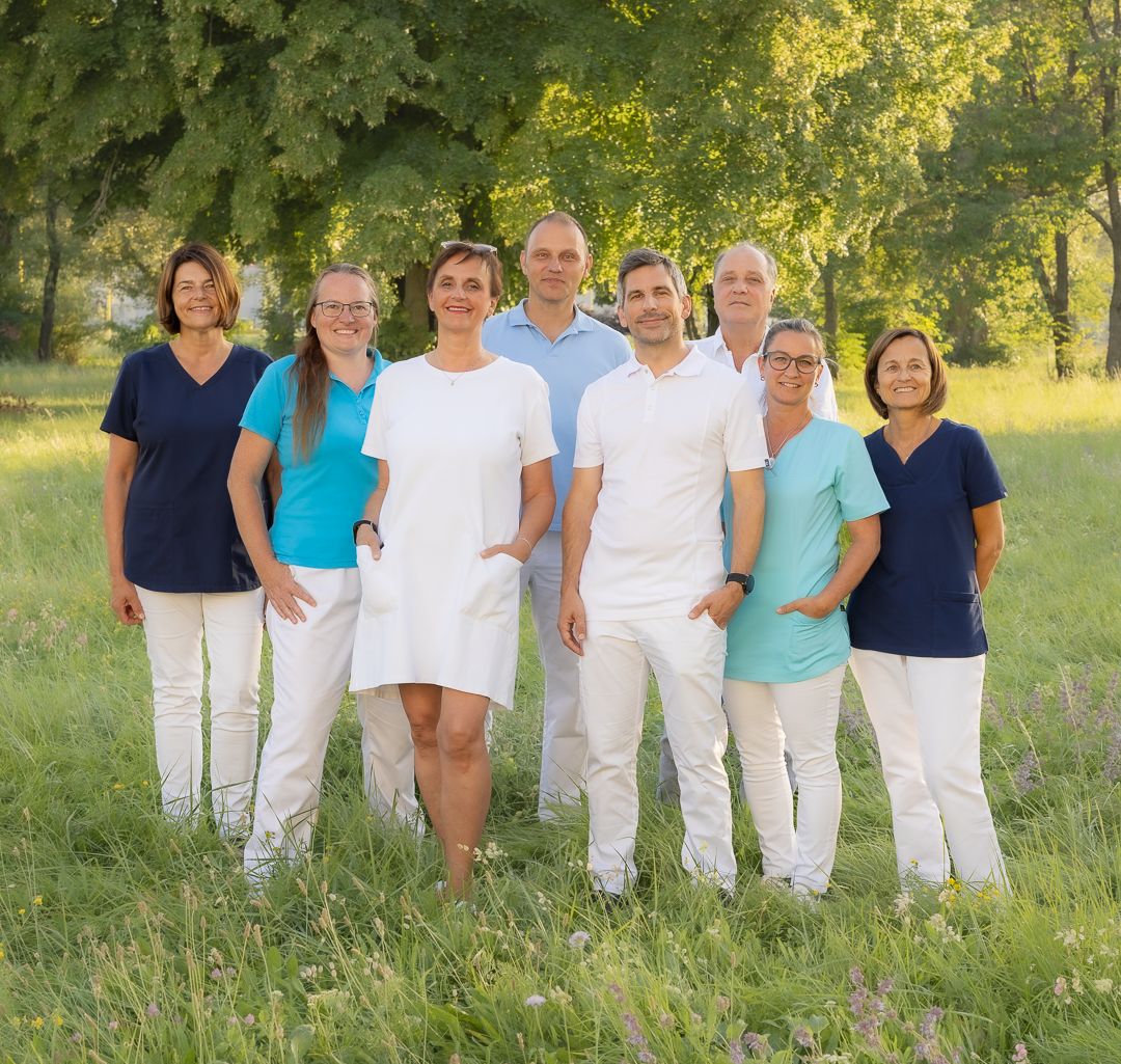 Gruppenfoto des Ortho-Vital-Teams: neun Personen in Praxiskleidung auf einer sommerlichen Wiese vor Bäumen